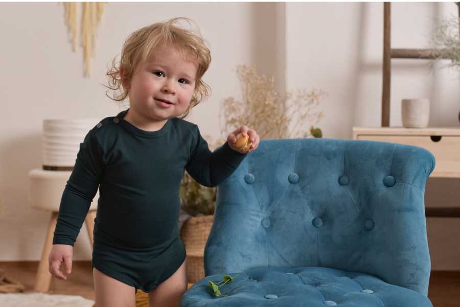 A blond curly boy of about one year old in a dark green bodysuit is standing by a blue chair
